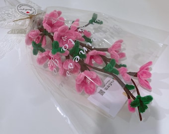 May include: A bouquet of pink and green pipe cleaner flowers in a clear plastic bag. The flowers are arranged in a vase-like shape and have a delicate, whimsical appearance.