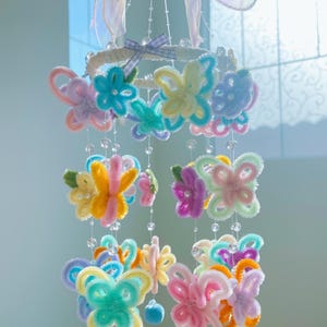 May include: A colorful mobile made of pipe cleaners shaped into butterflies and flowers. The mobile is hanging from a white hoop with a pink ribbon and bell. The mobile is decorated with clear beads.