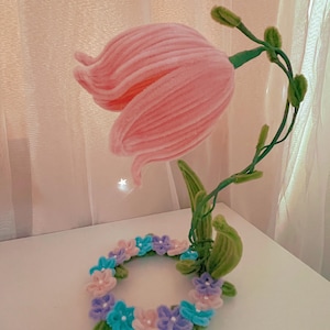 May include: A pink pipe cleaner flower with green stems and leaves, surrounded by a wreath of smaller pipe cleaner flowers in pink, blue, and purple.