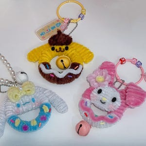 May include: Three colorful donut-shaped plush keychains with bells. The keychains are decorated with sprinkles and have different character faces. One is yellow with a brown face, one is pink with a white face, and one is white with a blue donut.