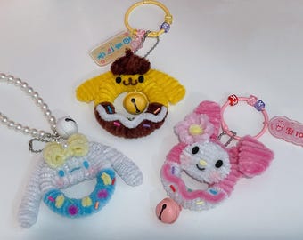 May include: Three colorful donut-shaped plush keychains with bells. The keychains are decorated with sprinkles and have different character faces. One is yellow with a brown face, one is pink with a white face, and one is white with a blue donut.