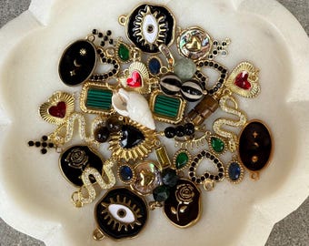 May include: A collection of assorted jewelry charms in gold-tone metal, featuring various shapes including hearts, eyes, roses, and snakes. The charms are decorated with black, white, red, green, and blue enamel, clear crystals, and a small white seashell.