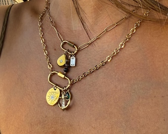 May include: A layered gold-tone necklace set featuring two distinct chains connected by oval carabiner clasps. The charms include a sunburst disc with a clear stone, a small anchor with a green gem, a teardrop charm, and a rectangular crystal.