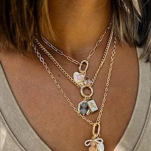 Puede incluir: Un conjunto de collares dorados en capas con varios colgantes. El collar superior presenta un dije de corazón rosa, el del medio un signo zodiacal y el inferior un colgante de lazo y perla. Las cadenas tienen diferentes estilos.
