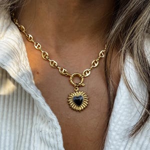 May include: Gold-toned necklace featuring a heart-shaped pendant with a black center and sunburst design. The chain has a link design and a circular clasp. The necklace is worn against a white shirt.