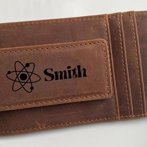 May include: A brown leather wallet with a magnetic money clip. The clip features a black atom symbol and the name "Smith". The wallet has multiple card slots and a stitched border.