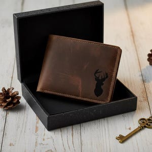 May include: A brown leather wallet with a black deer silhouette is displayed inside a black gift box. The wallet has a textured appearance and is rectangular. The box is open, revealing the wallet. A pine cone and a decorative key are also visible.