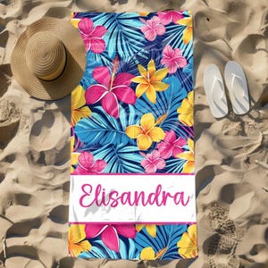 Pode incluir: Uma toalha de praia com um padr&atilde;o floral vibrante com flores e folhas rosa, amarelas e azuis. A toalha tem o nome "Elisandra" impresso em rosa numa faixa retangular branca. Um chap&eacute;u de palha e chinelos brancos est&atilde;o ao lado.