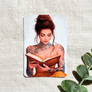 May include: A rectangular sticker featuring an illustration of a person with auburn hair in an updo, wearing a yellow strapless top. The person has visible tattoos on their neck, chest, and arms, and is holding an open brown book while looking down.