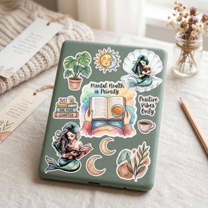 A sage green e-reader decorated with bookish stickers on a linen surface with a bookmark, dried flowers, and a pencil. Stickers include two teal mermaids reading, a monstera plant, a sun and moon face, hands holding an open book with coffee, a crescent moon, and phrases "Mental Health is Priority," "Just One More Chapter," and "Positive Vibes Only."