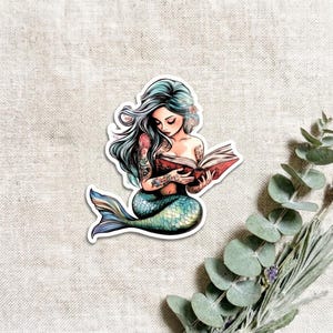 A single teal mermaid vinyl sticker on a linen background beside fresh eucalyptus sprigs. The mermaid has long flowing silver-blue hair and a green-blue tail, reading an open book.