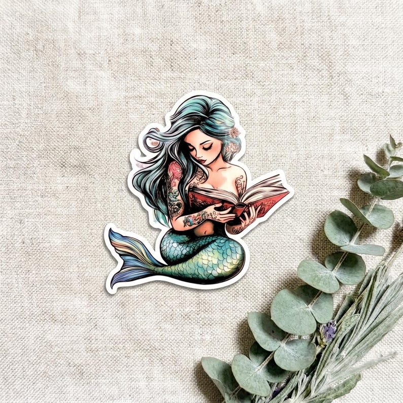 A single teal mermaid vinyl sticker on a linen background beside fresh eucalyptus sprigs. The mermaid has long flowing silver-blue hair and a green-blue tail, reading an open book.
