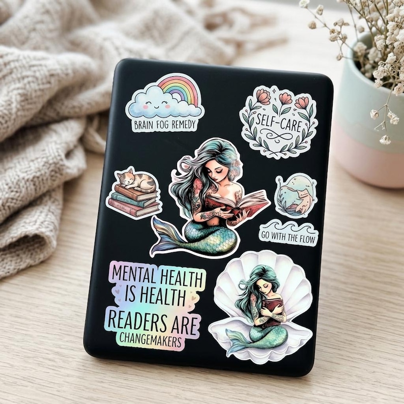 A black e-reader covered in bookish mental health stickers, resting on a knit blanket beside baby's breath flowers. Stickers include two teal mermaids reading, a cat napping on books, a rainbow cloud, and phrases "Brain Fog Remedy," "Self-Care," "Go with the Flow," "Mental Health is Health," and "Readers are Changemakers."