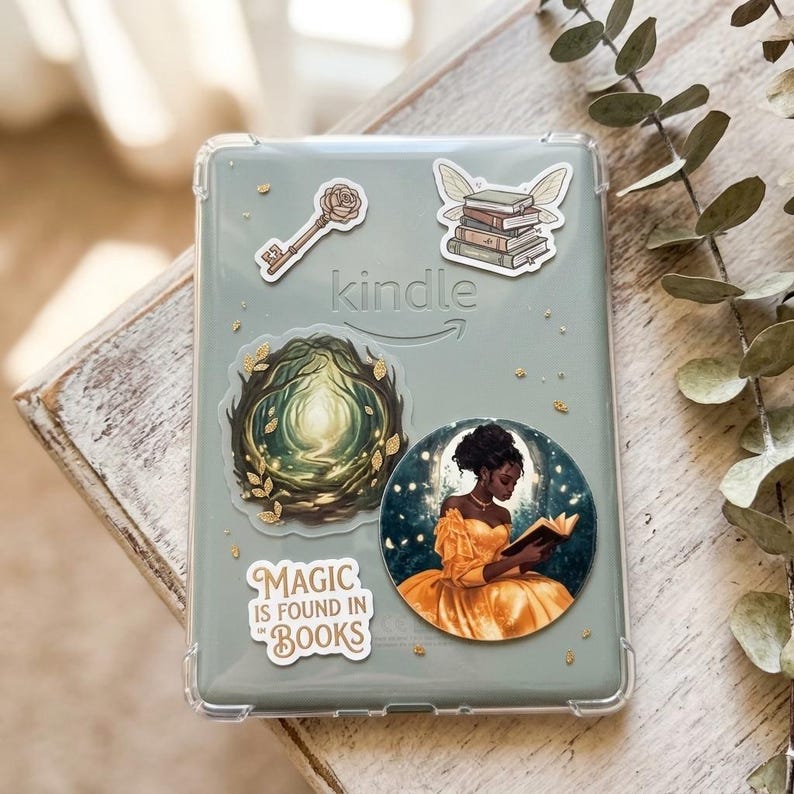 Clear Kindle e-reader case decorated with vinyl stickers, including a rose-topped key, a stack of books with butterfly wings, a glowing forest portal, a Magic is Found in Books quote, and a Black woman in a yellow gown reading inside a glowing moon, with gold accents and eucalyptus on a rustic white wood surface.