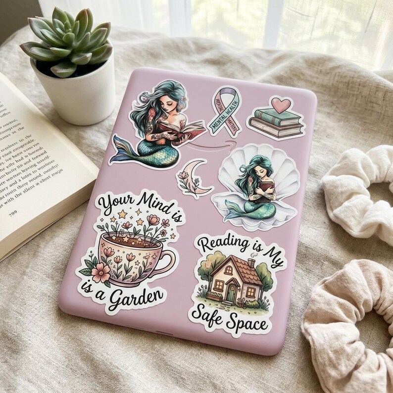 A lavender Kindle e-reader case decorated with the bookish sticker set, beside a potted succulent and an open book. Stickers include two teal mermaids reading, a Mental Health Matters ribbon, stacked books with a heart, a floral teacup reading "Your Mind is a Garden," and a cottage reading "Reading is My Safe Space."