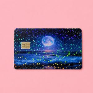 May include: A credit card with a holographic design featuring a full moon over a dark blue ocean with sparkling accents. The card has a gold-colored chip on the left side and is set against a pink background.