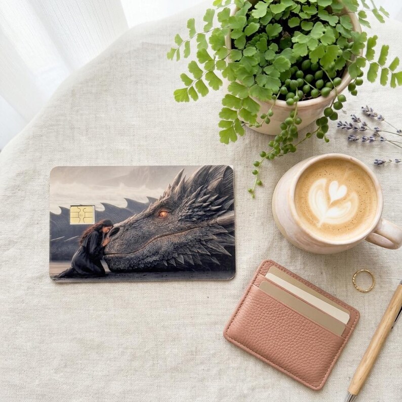 a debit card skin of a black dragon laying it's head down so a human can rest her head on the dragon on a cream colored cloth, a small green plant and a pinkish colored wallet on the side. 
and a hot coffee
 made by thestickerera