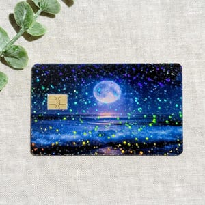 May include: A rectangular credit card skin featuring a dark night sky and ocean scene with a full moon. The surface has a holographic glitter finish with scattered green and blue light reflections. A gold metallic chip is visible on the left side.