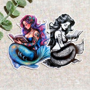 以下が含まれることがあります： 本を読む人魚が描かれた2枚のダイカットステッカー。1枚はピンクと紫の長い髪、青い尾、腕にカラフルなタトゥーがある人魚。もう1枚は、長い髪と腕にタトゥーがある人魚の白黒イラストです。