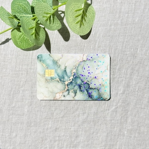 May include: A rectangular payment card featuring a marbled pattern in shades of white, sage green, and deep teal, accented with gold veining and scattered iridescent purple glitter. A gold-colored EMV chip is embedded on the left side of the card.