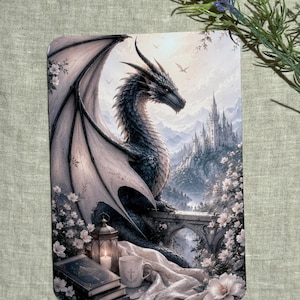 May include: A rectangular print featuring a dark, scaled dragon perched on a stone bridge near a castle. In the foreground, a lit lantern, a stack of books, and a white mug sit on a draped cloth surrounded by white flowers under a soft sky.