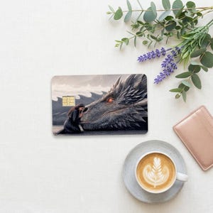 a debit card skin of a black dragon laying it's head down so a human can rest her head on the dragon on a cream colored cloth, flower and a hot coffee
 made by thestickerera