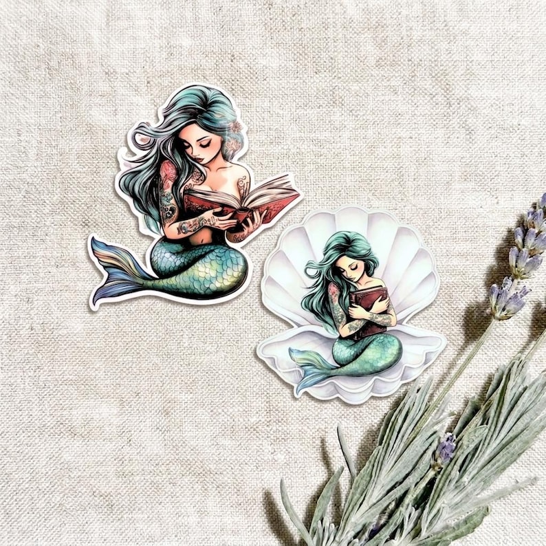 Two teal mermaid vinyl stickers on a linen background with lavender sprigs. One mermaid with flowing silver hair reads an open book; the other sits inside a white seashell holding a book, both with green-blue tails.
