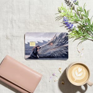 a debit card skin of a black dragon laying it's head down so a human can rest her head on the dragon on a cream colored cloth, a lavander 
plant and a pinkish colored wallet on the side. 
and a hot coffee
 made by thestickerera
