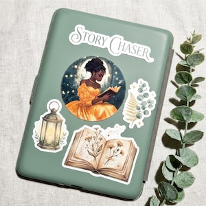 Sage green e-reader case decorated with vinyl stickers, including a Story Chaser banner, a Black woman in a yellow gown reading inside a glowing moon, a glowing lantern, an open book with pressed wildflowers, and a fern, with eucalyptus leaves on the side.
