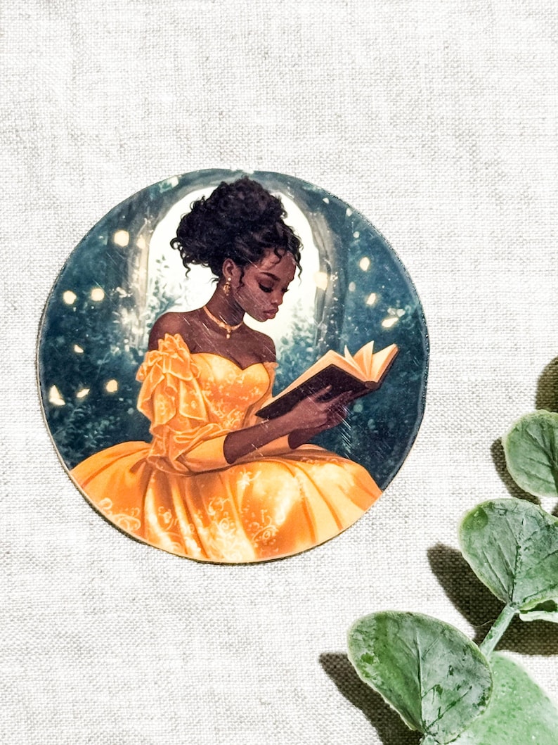 Vinyl sticker of a Black woman in a flowing yellow off-shoulder gown reading a book inside a glowing circular moon, surrounded by fireflies, set against a linen background with green eucalyptus leaves.