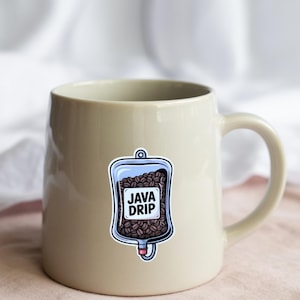Op de afbeelding: Een beige keramische mok met een sticker van een blauwe IV-zak gevuld met koffiebonen. De sticker heeft de woorden "JAVA DRIP" in witte blokletters. De mok staat op een roze ondergrond.