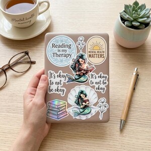 A hand holds a tan e-reader decorated with bookish mental health stickers, next to a coffee cup, glasses, a pen, and a succulent. Stickers include two teal mermaids reading, holographic stacked books, and phrases "Reading is my Therapy," "Mental Health Matters," and "It's okay to not be okay."