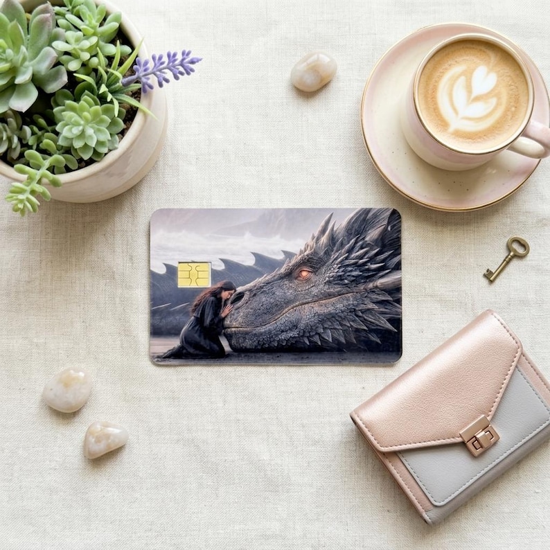 a debit card skin of a black dragon laying it's head down so a human can rest her head on the dragon on a cream colored cloth, a small green plant and a pinkish colored wallet on the side. 
and a hot coffee with a small key on the side. 

 made by thestickerera