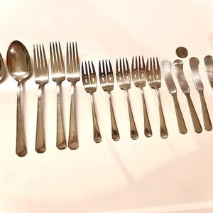 Könnte beinhalten: Ein Besteckset aus poliertem Silber, bestehend aus Gabeln, Löffeln und Buttermessern. Das Besteck ist in einer Reihe auf einer weißen Oberfläche angeordnet.