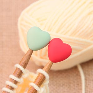 May include: Two wooden knitting needles with heart-shaped toppers, one mint green and one red, are shown. The needles are inserted into a piece of knitted yarn, with a large ball of yellow yarn in the background. The needles are a craft supply.
