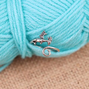 May include: A silver-colored dragon ring with a detailed design. The dragon has wings and is wrapped around a finger. The ring is set against a backdrop of a large ball of light blue yarn.