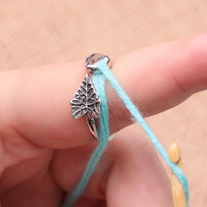 Op de afbeelding: Een zilverkleurige ring in de vorm van een blad met lichtblauw garen. De ring zit om een vinger, met een haaknaald op de achtergrond. Het garen is een heldere, effen kleur, ideaal voor haken.