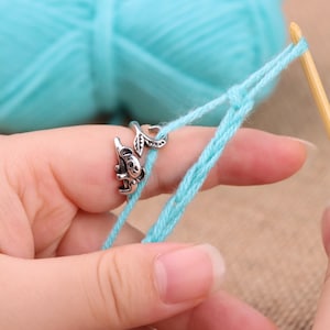 Puede incluir: Anillo guía de hilo plateado con diseño de koala y hoja, utilizado para tejer o hacer ganchillo. El anillo está en un dedo, con hilo azul claro que se está trabajando. Una madeja de hilo azul claro está en el fondo.