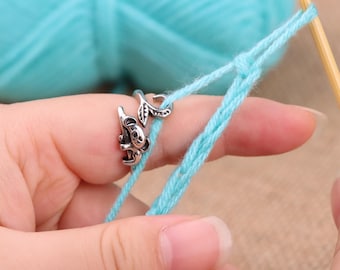 Peut inclure: Anneau guide-fil argenté avec un motif de koala et de feuille, utilisé pour le tricot ou le crochet. L'anneau est sur un doigt, avec du fil bleu clair en cours de travail. Une pelote de fil bleu clair est en arrière-plan.