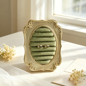 May include: An ornate, oval-shaped ring holder with a sage green velvet interior. The frame is a light, antique gold color. Two gold rings are nestled in the velvet. The holder sits on a white surface, with flowers and a card nearby.