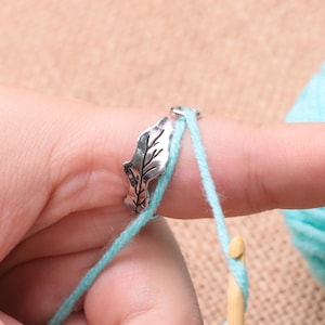 May include: A silver leaf-shaped yarn guide ring is on a finger, with light blue yarn threaded through it. A wooden crochet hook is in the foreground, with a ball of light blue yarn in the background. The image is a close-up.