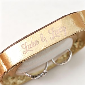 K&ouml;nnte beinhalten: Eine goldfarbene Schmuckschatulle aus Metall in einer geschwungenen, l&auml;nglichen Ovalform. Der Deckel ist mit dem eingravierten Text "Luke & Lucy" in Schreibschrift versehen. Das Innere enth&auml;lt ein helles, weiches Kissen mit zwei Silberringen.