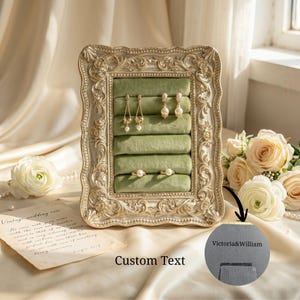 May include: A rectangular jewellery display frame with an ornate, cream-coloured, carved border and a sage green velvet interior featuring five horizontal slots. The display holds two pairs of pearl drop earrings and two pearl rings. Text reads Victoria&William.