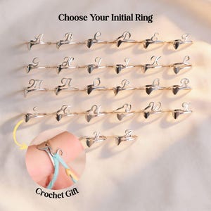 Puede incluir: Anillos iniciales plateados, cada uno con la forma de una letra del alfabeto, se muestran. Los anillos están sobre un fondo claro. El texto "Choose Your Initial Ring" está en la parte superior. Un primer plano muestra un anillo que se utiliza como regalo de ganchillo.