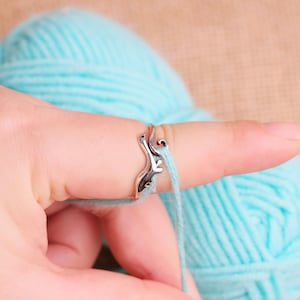 May include: A silver-colored yarn guide ring with a decorative design is worn on a finger, with light blue yarn passing through it. A ball of light blue yarn is in the background. The ring is designed to help with knitting or crocheting.