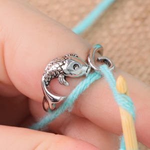 May include: A silver-colored fish-shaped yarn guide ring. The ring is holding a strand of light blue yarn, with a wooden crochet hook in the background. The fish has detailed scales and fins.