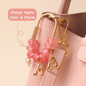May include: A gold-toned metal carabiner bag charm featuring pink agate beads, a small white pearl, and two gold-toned letter charms, M and A. The charm is attached to a textured pink handbag. Text in the image reads: Choose Agate Color & Charm.