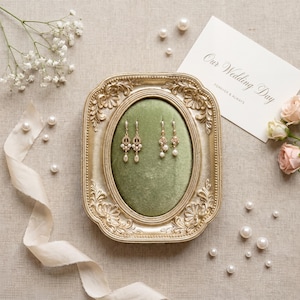 K&ouml;nnte beinhalten: Ein Paar goldfarbene Ohrh&auml;nger mit kleinen wei&szlig;en Perlen, pr&auml;sentiert auf einem gr&uuml;nen Samtkissen in einem verzierten, rechteckigen Goldrahmen. Eine wei&szlig;e Karte mit dem Text Our Wedding Day liegt neben dem Rahmen.