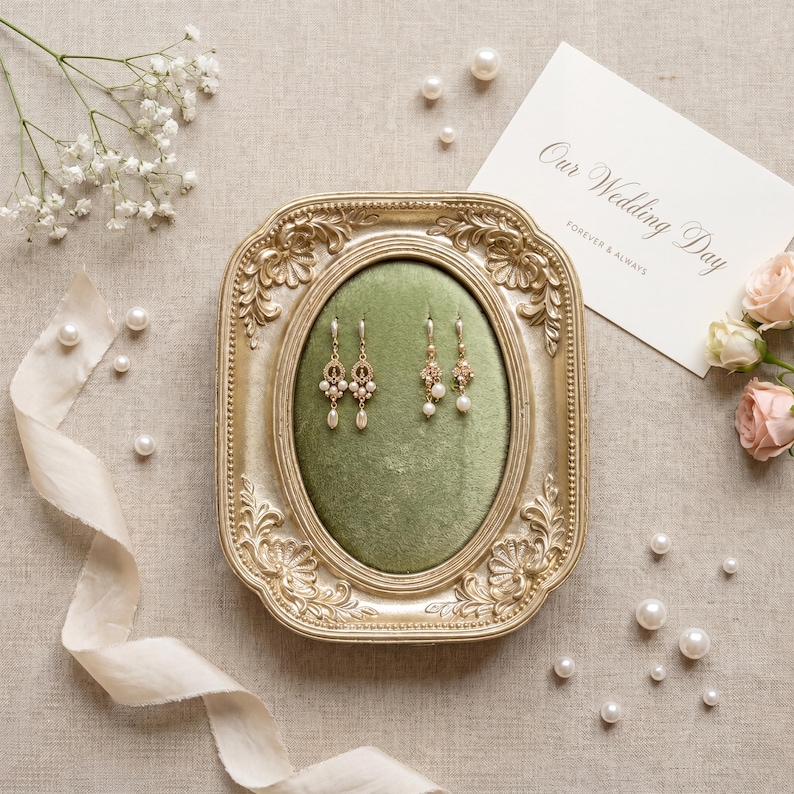 K&ouml;nnte beinhalten: Ein Paar goldfarbene Ohrh&auml;nger mit kleinen wei&szlig;en Perlen, pr&auml;sentiert auf einem gr&uuml;nen Samtkissen in einem verzierten, rechteckigen Goldrahmen. Eine wei&szlig;e Karte mit dem Text Our Wedding Day liegt neben dem Rahmen.