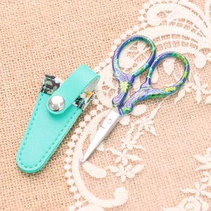 Puede incluir: Tijeras decorativas pequeñas con diseño floral en azul y verde, sobre una superficie beige. Incluyen una funda de polipiel turquesa con solapa estampada floral y cierre de botón plateado.